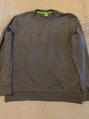Grå sweatshirt från BOSS - Snygg grå sweatshirt från BOSS med diskret broderad logga på bröstet. Tröjan har rund halsringning, långa ärmar och ribbade muddar vid ärmslut och nederkant. Perfekt för en avslappnad och stilren look.