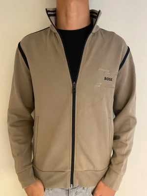 Beige zip hoodie från BOSS - Snygg beige hoodie från BOSS med hel dragkedja och svarta detaljer längs dragkedjan och axlarna. Ribbad krage med svarta ränder och diskret BOSS-logga på bröstet. Två fickor framtill och mjukt material som känns skönt mot huden.
