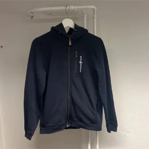 Mörkblå jacka från Sail Racing - Mörkblå jacka från Sail Racing med dragkedja framtill och en bröstficka med logga. Jackan har hög krage och sportig look, perfekt för kyliga dagar. Snygg och enkel design med praktiska detaljer.