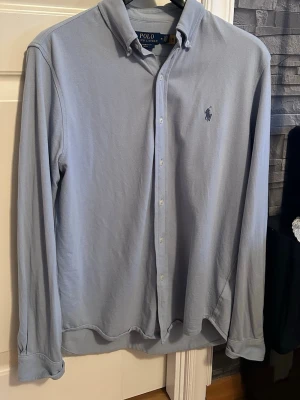Skjorta från Polo Ralph Lauren - Klassisk ljusblå skjorta från Polo Ralph Lauren med button-down krage och broderad logga på bröstet. Skjortan har långa ärmar och knäppning framtill. Tillverkad i mjuk bomull för en bekväm känsla.