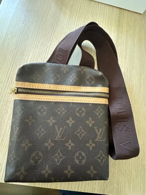 Brun axelväska med monogram - Snygg brun axelväska med klassiskt monogrammönster och justerbar bred axelrem i tyg. Väskan har guldfärgad dragkedja och detaljer i beige. Perfekt storlek för det viktigaste och en stilren accessoar till din outfit.