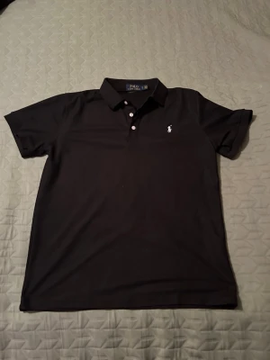 Svart pikétröja från Polo Ralph Lauren - Klassisk svart pikétröja från Polo Ralph Lauren med vit broderad logga på bröstet. Tröjan har korta ärmar, krage och knäppning med tre vita knappar framtill. Perfekt till jeans eller shorts för en clean och stilren look.