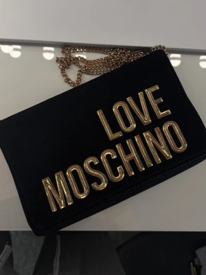 Axelväska från Love Moschino - Snygg svart axelväska från Love Moschino med stora guldfärgade bokstäver på framsidan och kedjerem i guld. Väskan är i ett mjukt material och har ett innerfack med logga. Dust bag medföljer.