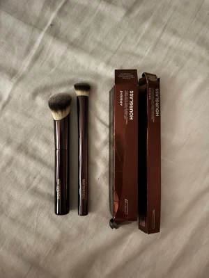 Hourglass sminkborstar duo - Två exklusiva sminkborstar från Hourglass med metalliskt mörkbruna handtag och täta, mjuka borst i svart och vitt. Perfekta för contouring och applicering av puder eller foundation. Snygg design och levereras med originalförpackning.