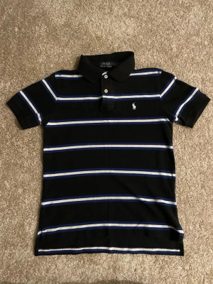 Svart pikétröja från Polo Ralph Lauren - Svart pikétröja från Polo Ralph Lauren med blå och vita horisontella ränder. Klassisk krage, två knappar och broderad logga på bröstet. Perfekt för en stilren och avslappnad look.