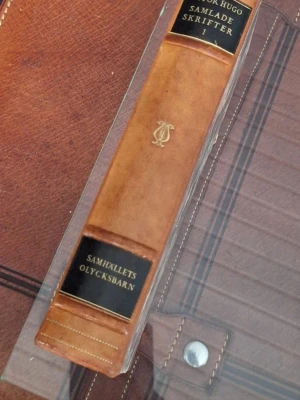 1927 Victor Hugo Samlade skrifter I - Säljer första delen av Victor Hugos samlade skrifter, 'Samhällets olycksbarn'. Boken har ett klassiskt brunt läderband med gulddetaljer och svarta etiketter på ryggen. Den är från 1927
