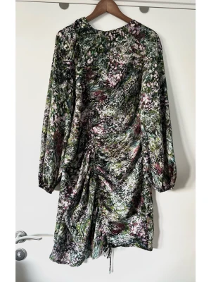 Blommig kortklänning med snörning - Härlig flerfärgad kort klänning från Vero Moda i storlek XL. Stilren modell med långa ärmar och rund halsringning. Klänningen har en justerbar dragsko tvärsöver framsidan som skapar en draperad effekt och en asymmetrisk fåll. Mönstret är abstrakt med inslag av grönt, lila, rosa och svart. Diskret nyckelhålsöppning med knapp baktill i nacken och en praktisk dold dragkedja i sidan. Halvstretchigt mesh-material för bästa passform. Smickrande modell på alla kroppsformer. Mycket gott skick, som ny!