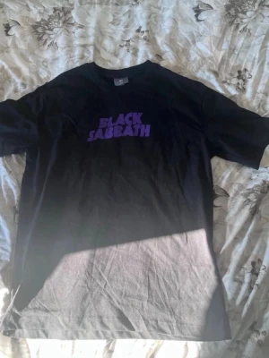 Svart Black Sabbath t-shirt - Säljer en svart t-shirt med lila 'Black Sabbath'-tryck på bröstet. Klassisk rund hals och korta ärmar. Perfekt för dig som gillar bandmerch och vill ha en stilren men ändå uttrycksfull look.