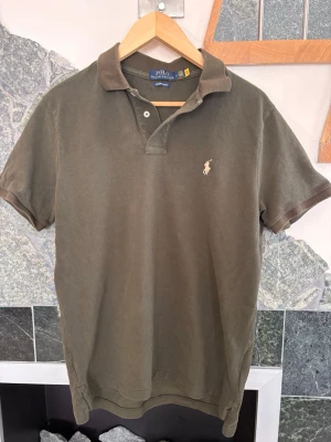 Olivgrön pikétröja från Polo Ralph Lauren - Klassisk olivgrön pikétröja med korta ärmar och krage. Tröjan har två knappar vid halsen och den ikoniska broderade loggan i vitt på bröstet. Perfekt för en avslappnad men ändå stilren look.