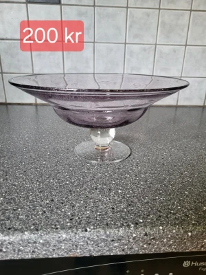 Lila glasskål på fot - Säljer en snygg lila glasskål på fot med bred kant och rundad form. Skålen har en genomskinlig fot och passar perfekt som fruktfat eller dekoration. Materialet är glas och färgen är en diskret lila ton.
