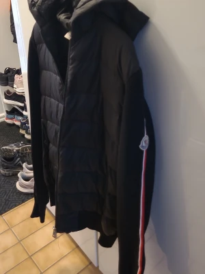Moncler cardigan - Snygg svart dunjacka med huva och quiltad design. Jackan har en röd och vit rand längs ärmen samt en patch på överarmen. Perfekt för kalla vinterdagar och har en sportig look som passar till det mesta. Står att det är storlek L men Passar även s-m.den är knappt använd men Det är ett hål I armhålan men det är lättfixat då det är precis vid en sum, kan även fixa de innan jag postar om man vill de
