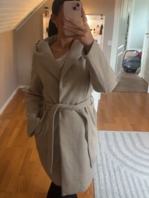 Beige kappa med knytskärp - Stilren beige kappa med luva och knytskärp i midjan. Kappan har en avslappnad passform, två sidofickor och är tillverkad i ett mjukt tyg som ger en lyxig känsla. Perfekt för kyligare dagar och enkel att matcha med olika outfits.