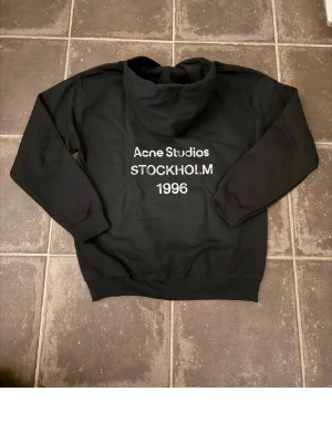 Svart hoodie Acne Studios Stockholm - Svart hoodie från Acne Studios med tryck 'Acne Studios STOCKHOLM 1996' på bröstet. Klassisk passform med huva och känguruficka framtill. Tillverkad i mjukt material, perfekt för en chill och stilren look. Jag är 185, vikt ≈ 68. Skriv för fler frågor osv.