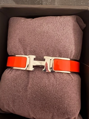Rött armband med H-spänne - Snyggt armband i röd lackad metall med ett stort H-format spänne i silverfärg. Armbandet har en stilren och modern design som sticker ut och passar perfekt till dig som gillar statement-accessoarer.