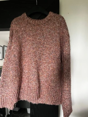 Rosa melerad stickad tröja Indiska - Mysig stickad tröja från Indiska i en rosa melerad färg med inslag av lila och orange. Tröjan har rund halsringning, långa ärmar och ribbade muddar vid ärmslut och nederkant. Perfekt för kyliga dagar och enkel att matcha med jeans eller kjol.