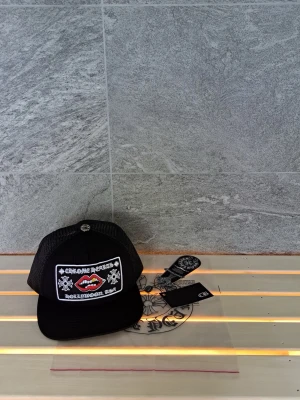 Svart Chrome Hearts keps med nät - Säljer en svart keps från Chrome Hearts med mesh på sidorna och bak, samt ett broderat märke framtill med vita detaljer och röda läppar. Kepsen har justerbar snapback och cool metallknapp på toppen. Perfekt för dig som gillar streetwear och unika accessoarer.