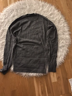 Mörkgrå merinoull tröja - En enkel och stilren mörkgrå stickad tröja med rund halsringning och långa ärmar. Tröjan är tunnstickad och har ribbade muddar vid hals, ärmslut och nederkant. Perfekt för lager på lager under kyliga dagar.