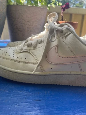 Vita låga nike court med rosa swoosh stl 36 - Snygga vita sneakers i läder med klassisk swoosh på sidan. Skorna har perforerad tå för extra ventilation, vita skosnören och en låg siluett som ger en clean och tidlös look. Perfekta för dig som gillar streetwear och enkel stil.
