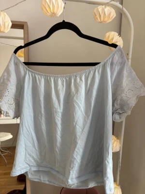 Ljusblå offshoulder topp med spets - Söt ljusblå offshoulder topp med korta ärmar och broderade spetsdetaljer. Toppen har en avslappnad passform och är perfekt för varma dagar. Tillverkad i mjukt material som känns skön mot huden.