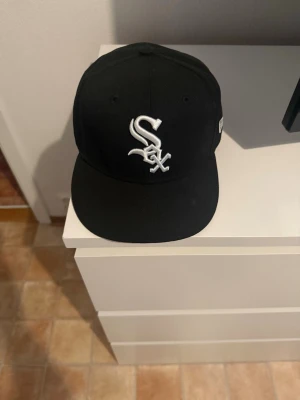 Svart keps med vit Sox-logga - Snygg svart keps med rak skärm och broderad vit Sox-logga framtill. Klassisk 59FIFTY-modell med insida i svart och grått. Perfekt för dig som gillar streetwear och sportig stil.