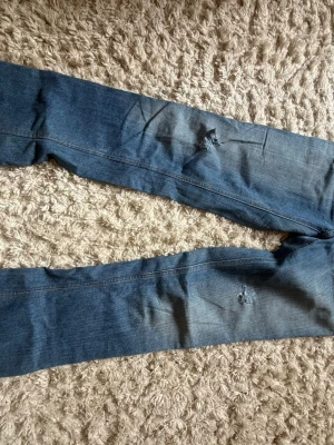 Blå slitna jeans med hål - Ett par blå jeans med smal passform och slitningar på benen. Jeansen har hål på framsidan och klassisk femficksdesign. Materialet är denim i bomull och färgen är mellanblå med viss tvättad effekt.