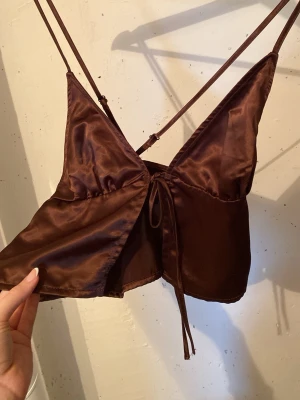 Brun satin linnetopp med knyt - Snygg brun linnetopp i satin med tunna axelband och knyt framtill. Toppen har en djup v-ringning och croppad passform, perfekt för sommarens varma dagar. Justerbara axelband för bästa passform.