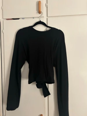 Svart långärmad topp med knyt - Säljer en svart långärmad topp med knyt i midjan. Toppen har en enkel, stilren design med rund halsringning och vida ärmar. Perfekt att styla med både jeans och kjol för en trendig look.