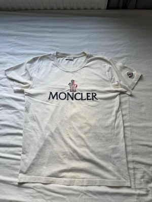 Vit  t-shirt  - Snygg vit t-shirt från Moncler med stor logga tryckt på bröstet och Moncler-märke på ena ärmen. Klassisk rund halsringning och korta ärmar. Tillverkad i mjuk bomull för en skön känsla och enkel stil.