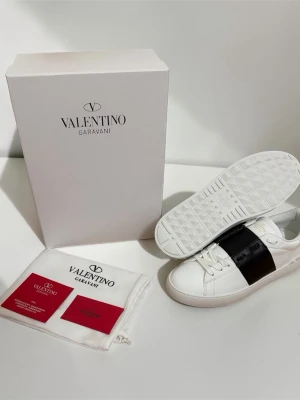 Valentino Garavani vita sneakers - Stilrena vita sneakers från Valentino Garavani med bred svart läderdetalj över mitten. Klassisk låg modell med vita snören och diskret logga på innersulan. Tillverkade i läder för en lyxig känsla och look.