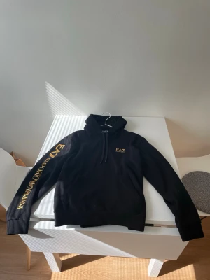 Svart hoodie från EA7 Emporio Armani - Snygg svart hoodie från EA7 Emporio Armani. Har guldfärgade logotexter på vänster ärm och bröstet. Klassisk passform med dragsko i huvan och ribbade muddar. Perfekt för en trendig och avslappnad stil.