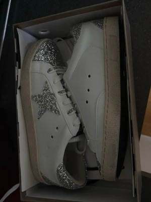 Vita sneakers med silverglitter - Snygga vita sneakers med silverglittrande detaljer på tå, häl och en stjärna på sidan. Skorna har vita snören och en beige sula. Perfekta för dig som vill sticka ut med lite extra bling i vardagen. Tillverkade i läder och syntetmaterial.