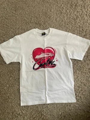 Cortiez Vit t-shirt med rött hjärta och kryssningsbåt - Vit t-shirt i bomull med tryck av ett stort rött hjärta och en kryssningsbåt på bröstet. Snygg grafisk text under motivet och klassisk passform med korta ärmar och rund hals. Perfekt för dig som gillar unika tryck och cruise-vibes.