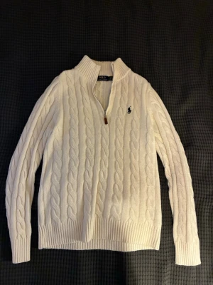 Vit kabelstickad tröja Polo Ralph Lauren - Kabelstickad vit tröja från Polo Ralph Lauren med halv dragkedja och hög krage. Tröjan har långa ärmar, ribbade muddar och den klassiska broderade loggan på bröstet. Perfekt för en clean och stilren look.