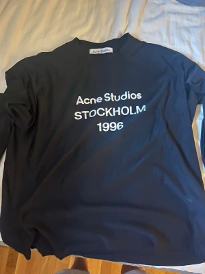 Svart Acne Studios longsleeve - Säljer en svart t-shirt från Acne Studios med vit text 'Acne Studios STOCKHOLM 1996' på bröstet. Tillverkad i 100% bomull och har en klassisk passform med korta ärmar och rund hals. Perfekt för dig som gillar stilrena och trendiga plagg.