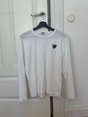 Vit långärmad topp Comme des Garçons - Säljer en vit långärmad topp från Comme des Garçons PLAY med det ikoniska svarta hjärtat broderat på bröstet. Enkel och clean design som passar till allt. Tillverkad i mjuk bomull för skön känsla hela dagen.
