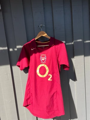 Arsenal matchtröja Henry 14 röd - Säljer en röd Arsenal matchtröja från Nike med guldfärgade detaljer, klubbmärke och O2-logga på bröstet. På ryggen står det HENRY och nummer 14 i guld, samt texten Highbury 1913-2006. Perfekt för dig som älskar fotboll och Arsenal.