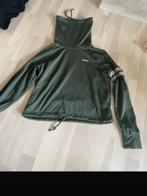 Grön hoodie från Stronger - Mörkgrön hoodie från Stronger med hög krage och dragsko i nederkant. Tröjan har långa ärmar och en vit logga på bröstet samt vita och svarta ränder på ena ärmen. Perfekt för träning eller chill.