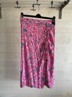 Blommig rosa kjol från Pieces - Säljer en blommig rosa kjol från Pieces i mjukt material. Kjolen har ett färgglatt mönster i blått, rött och grönt samt en snygg snörningsdetalj på sidan. Perfekt för dig som vill ha en färgklick i garderoben!