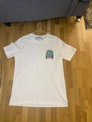 Vit Casablanca t-shirt med tryck - Säljer en vit t-shirt från Casablanca med färgglatt tryck på bröstet och stort grafiskt motiv på ryggen. T-shirten har rund hals och korta ärmar. Tillverkad i mjuk bomull för skön känsla och stilren look.