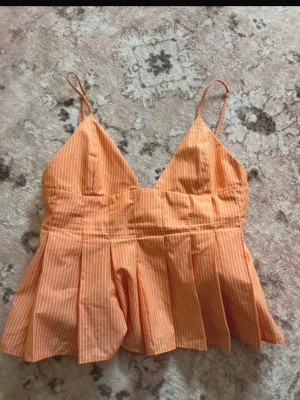 Randig orange linnetopp med tunna band - Söt randig linnetopp i orange med tunna axelband och v-ringad hals. Toppen har en luftig, utställd passform med veckade detaljer nedtill. Perfekt för varma dagar och ger en fräsch sommarkänsla.