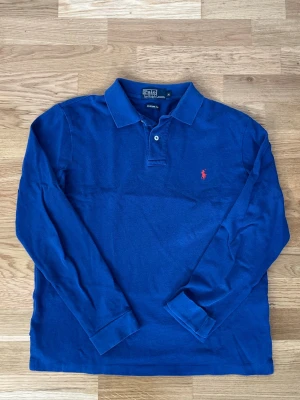 Blå långärmad pikéskjorta Polo Ralph Lauren - Snygg blå långärmad pikéskjorta från Polo Ralph Lauren med klassisk krage och knappar vid halsen. Ikoniska röda loggan broderad på bröstet. Perfekt för en stilren och avslappnad look.