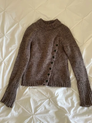 Brun stickad tröja med knappar - Mysig brunmelerad stickad tröja med asymmetrisk knappslå framtill. Tröjan har lång ärm med slits. Perfekt för kyliga dagar och enkel att styla till olika outfits.