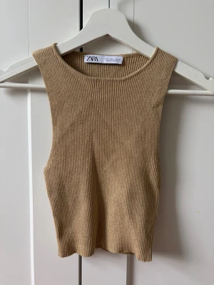 Guldig ribbad linnetopp från Zara - Säljer en guldig ribbad linnetopp från Zara. Toppen är ärmlös med rund halsringning och croppad passform. Perfekt att styla med höga jeans eller kjol för en trendig look.