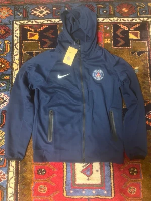 Mörkblå PSG träningsset från Nike - Snyggt mörkblått träningsset från Nike med PSG-logga på både jacka och byxor. Jackan har huva, hel dragkedja och två fickor med dragkedja. Byxorna har resår i midjan, dragkedjefickor och muddar vid benslut. Perfekt för fotbollsträning eller chill.
