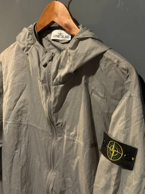 Grå vindjacka från Stone Island - Grå vindjacka från Stone Island med huva och dragkedja framtill. Jackan har den ikoniska logotypen på vänster ärm och är tillverkad i ett lätt syntetmaterial. Perfekt för dig som gillar stilren och funktionell design.