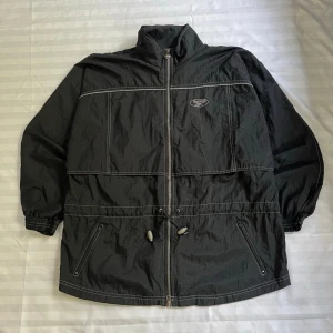 Svart vindjacka från Reebok - Svart vindjacka från Reebok med dragkedja framtill, hög krage och vita kontrastsömmar. Jackan har två snedställda fickor med dragkedja och justerbar dragsko i midjan. Tillverkad i lätt och tåligt syntetmaterial, perfekt för blåsiga dagar.