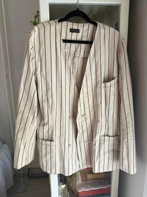 Randig beige kavaj med svarta ränder - Snygg beige kavaj med tunna svarta vertikala ränder. Dubbelknäppt modell med tre fickor framtill och avslappnad passform. Perfekt för en trendig och stilren look. 