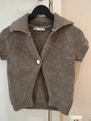 Trendig Beige stickad kortärmad kofta Zara - Kortärmad stickad kofta från Zara i beige melerad färg. Koftan har en stor krage, en knapp framtill och ribbade muddar vid ärmslut och nederkant. Perfekt att slänga över en topp för en chill look.