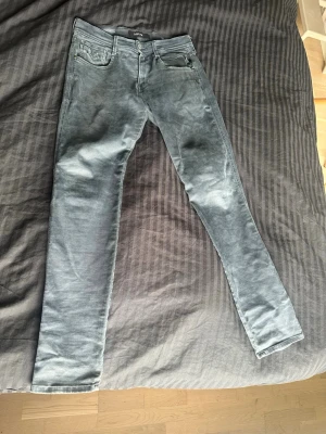 Replay Anbass Hyperflex - Säljer ett par gråa Replay jeans med klassisk femficksdesign och bälteshällor. Jeansen har en smal passform och är tillverkade i ett stretchigt denimtyg för extra komfort. Snygga detaljer på bakfickorna och en svart patch vid linningen.