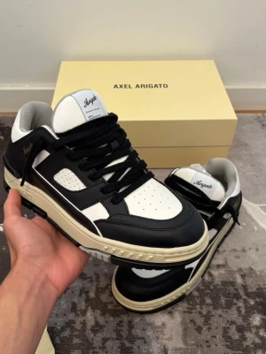 Oanvända Axel Arigato area lo - Snygga sneakers från Axel Arigato i svart och vitt läder med chunky sula och svarta skosnören. Klassisk low-top modell med gulddetaljer på sidan och perforerad tå för extra ventilation. Perfekta för dig som vill ha en clean och modern look.
